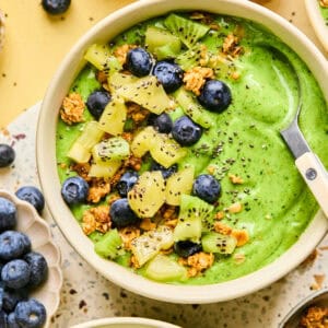 A vibrant smoothie bowl brims with lush green hues, adorned with sliced kiwi, blueberries, granola, and chia seeds. A spoon rests on the bowls edge. Whole kiwis and a small bowl of blueberries complete the scene on a speckled surface.