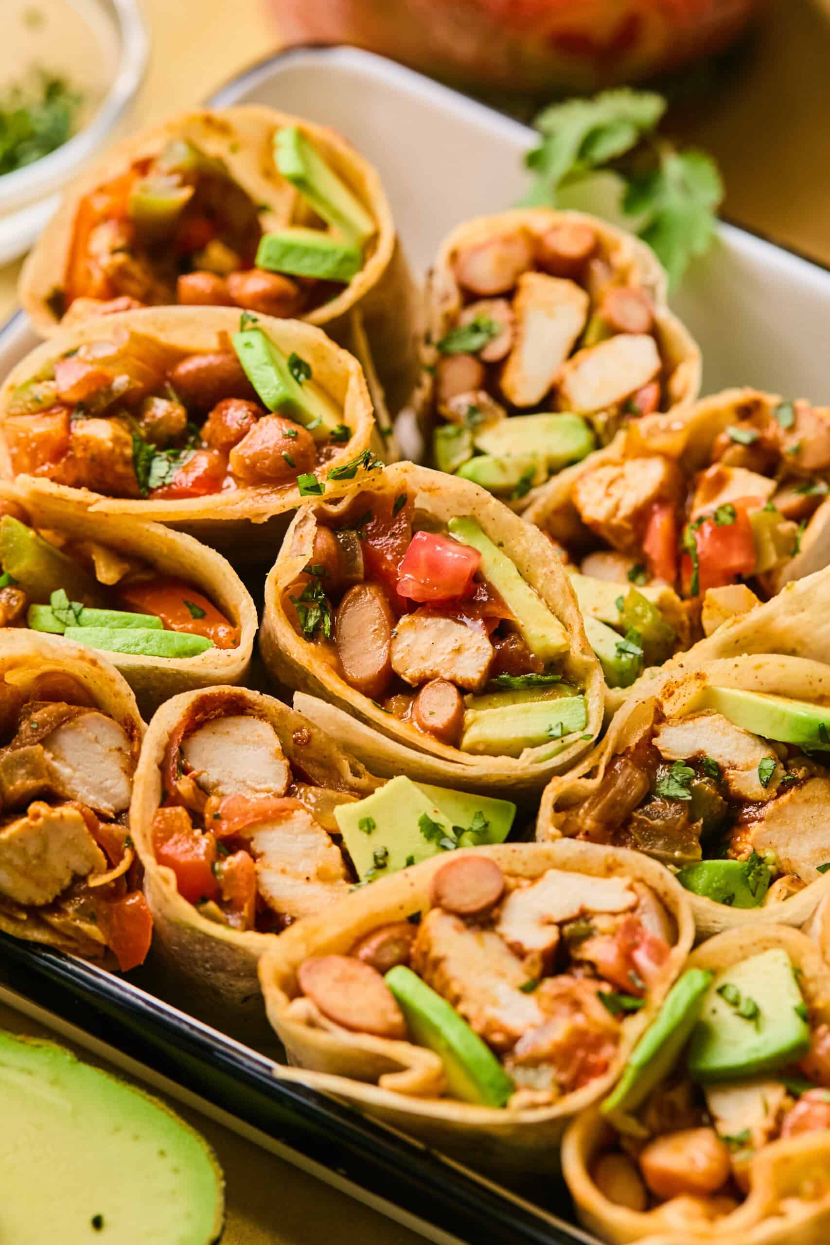 Close-up of a tray filled with assorted fajita wraps, each containing grilled chicken, diced tomatoes, beans, avocados, and fresh cilantro. The wraps are neatly arranged, showcasing vibrant colors and fresh ingredients, with an avocado half visible at the bottom.