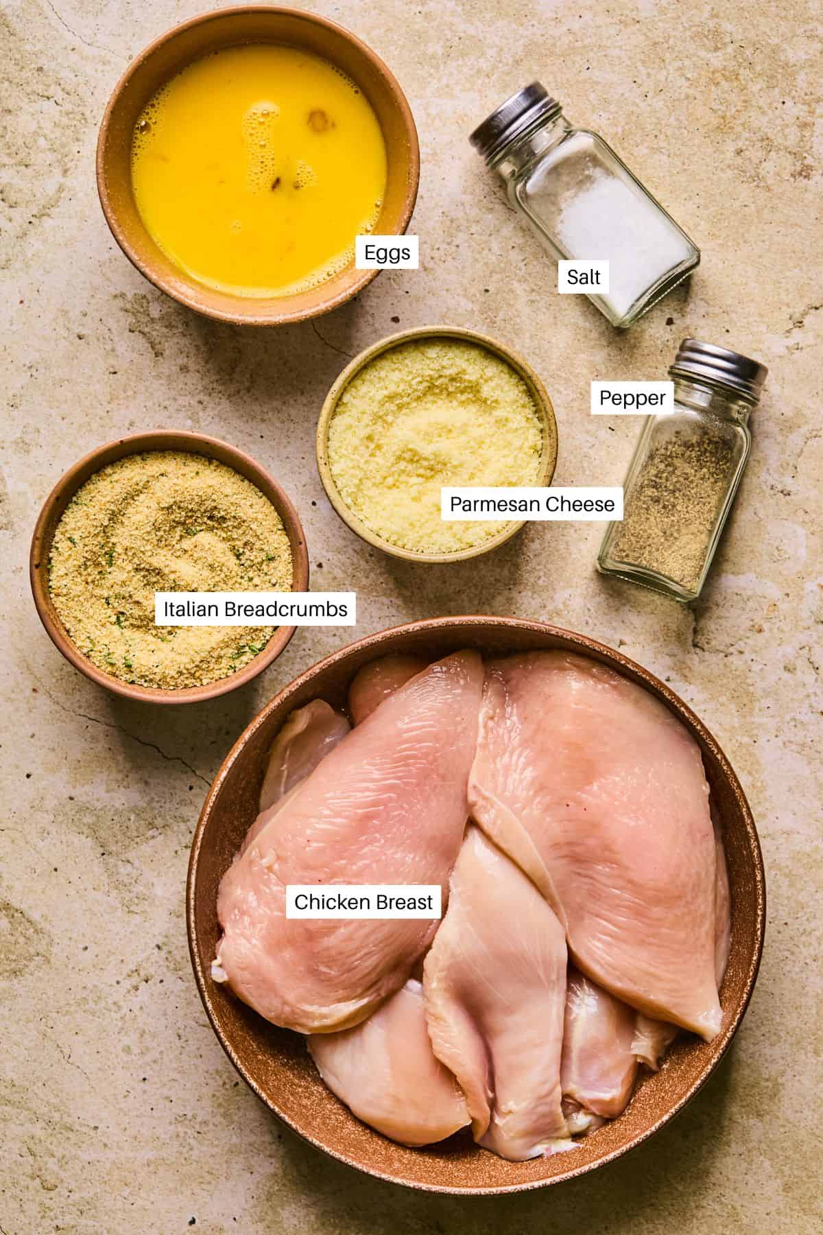 Top-down view of ingredients for baked chicken cutlets, featuring a bowl of raw chicken breasts, eggs, grated Parmesan cheese, Italian breadcrumbs, and shakers of salt and pepper, all artfully arranged on a light stone surface.