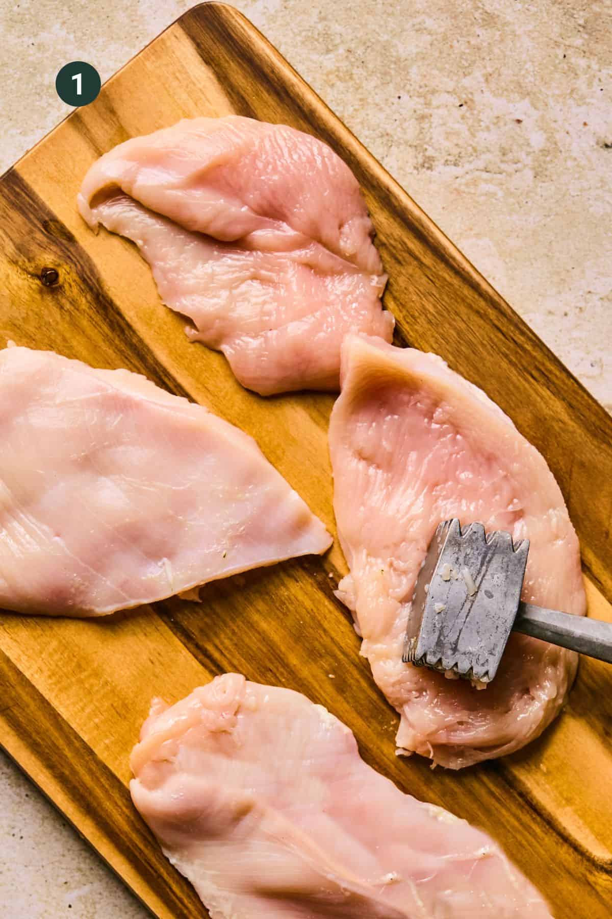 Four raw chicken breasts are being tenderized with a meat mallet on a wooden cutting board, ready to transform into delicious baked chicken cutlets. The board rests on a light textured surface, setting the stage for a delightful meal preparation.