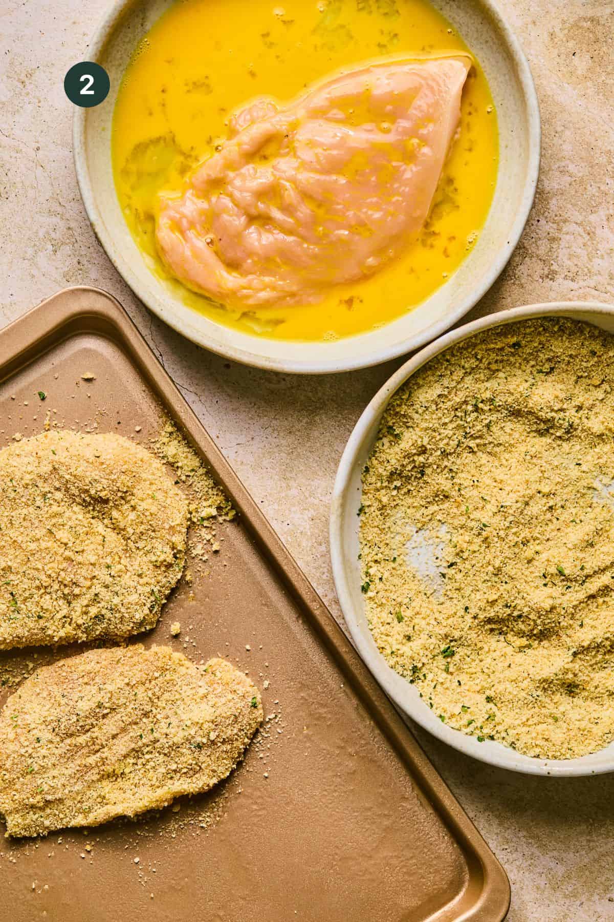 A chicken breast is being dipped in beaten egg in a bowl. Nearby, another bowl holds bread crumbs mixed with herbs. Two baked chicken cutlets are coated in the breadcrumb mixture on a baking sheet.