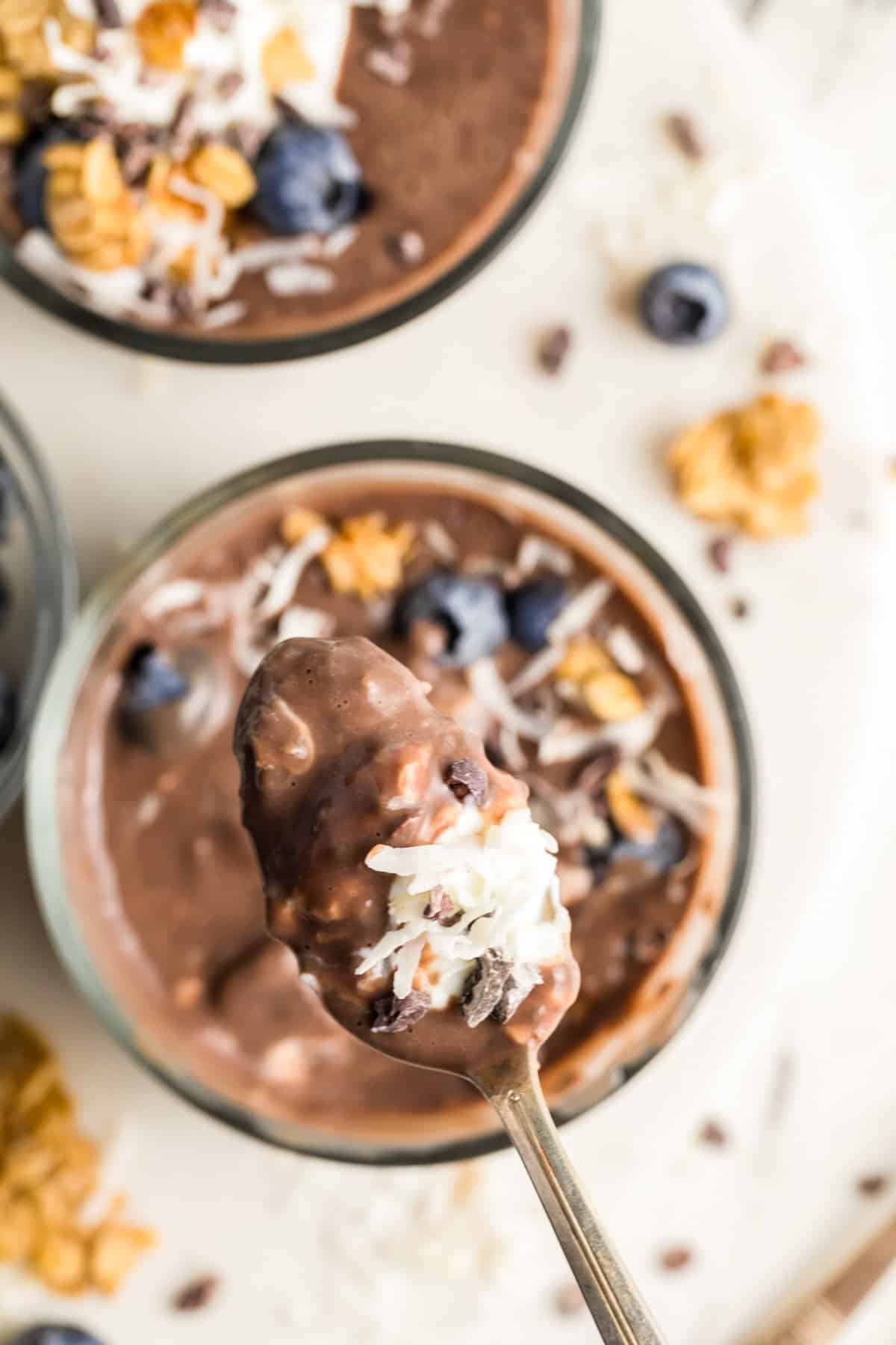 A spoonful of protein-packed creamy chocolate chia seed pudding topped with shredded coconut, blueberries, and granola is held above a bowl filled with the same delicious mix. Additional blueberries and granola are scattered around for an extra treat.