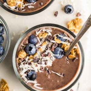 A bowl of chocolate smoothie topped with blueberries, granola, shredded coconut, and cacao nibs. A spoon is placed in the bowl alongside a sprinkle of protein-rich chia seed pudding, with more toppings scattered around on a marble surface.