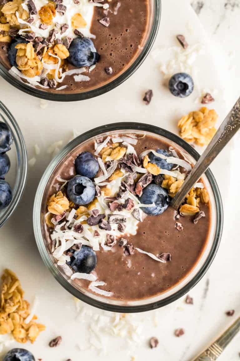 A bowl of chocolate smoothie topped with blueberries, granola, shredded coconut, and cacao nibs. A spoon is placed in the bowl alongside a sprinkle of protein-rich chia seed pudding, with more toppings scattered around on a marble surface.