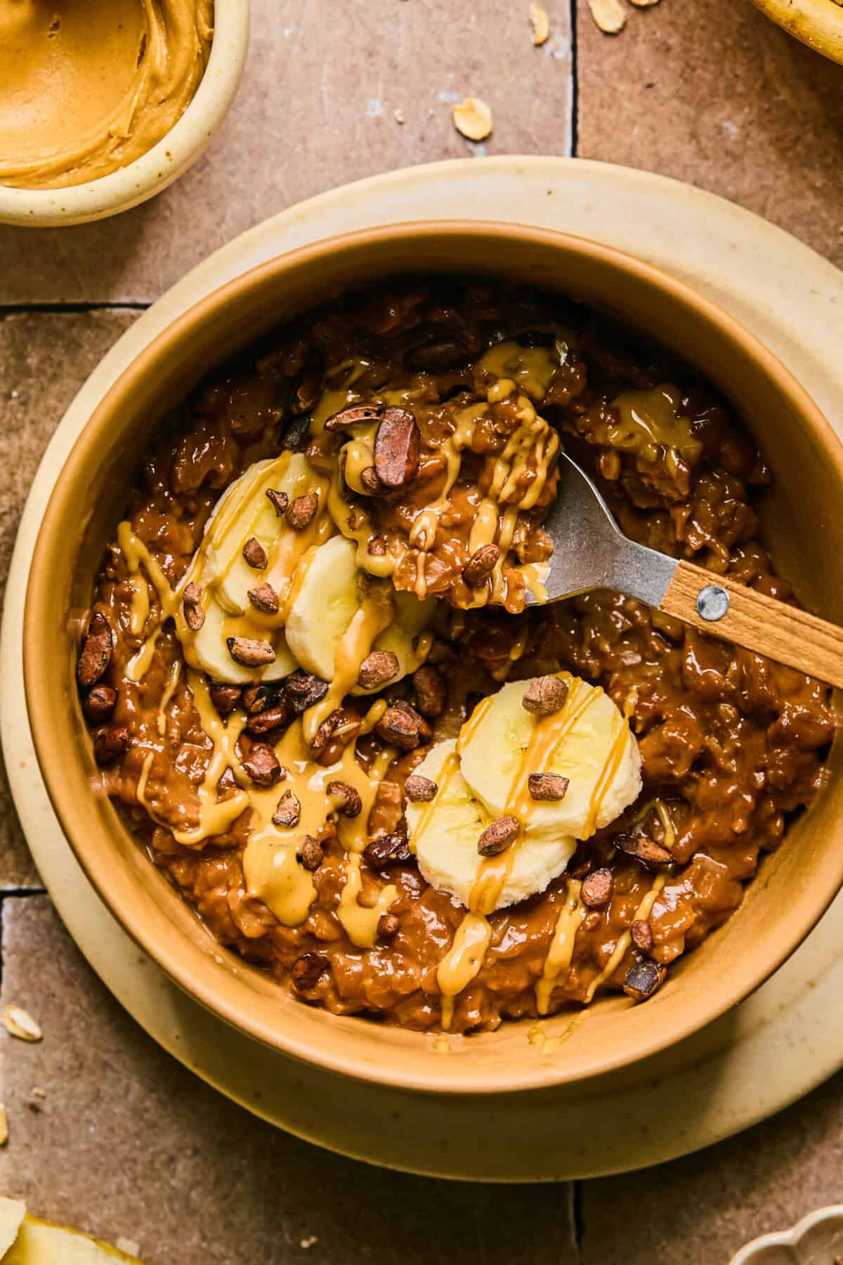 A bowl of chocolate protein oatmeal topped with banana slices, chocolate chips, and drizzled with creamy peanut butter. A wooden spoon rests inside the bowl. Surrounding the bowl are oats and small dishes on a tiled surface.