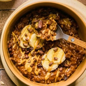 A bowl of chocolate protein oatmeal topped with banana slices, chocolate chips, and drizzled with creamy peanut butter. A wooden spoon rests inside the bowl. Surrounding the bowl are oats and small dishes on a tiled surface.