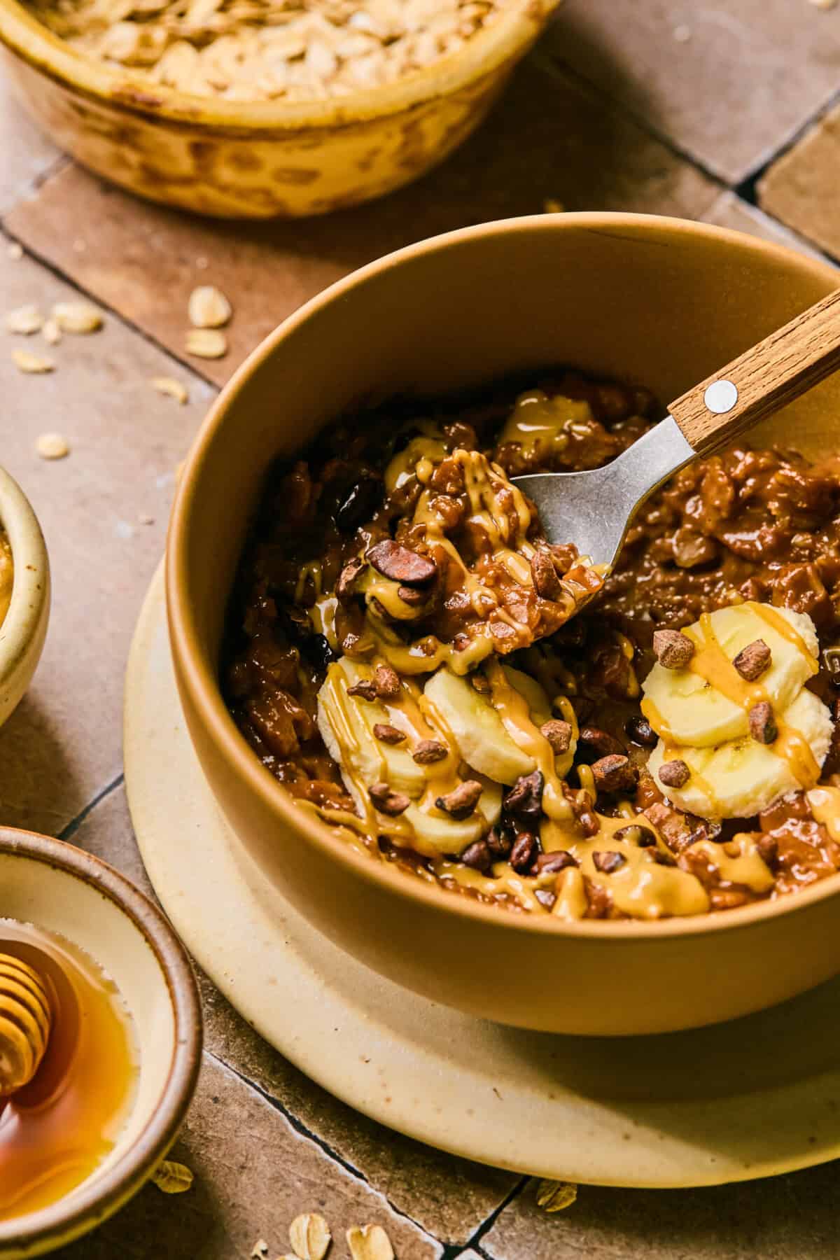 A bowl of protein oatmeal topped with banana slices, peanut butter, and cacao nibs, accompanied by a small wooden spoon. Nearby are additional bowls of honey and oats on a tiled surface.