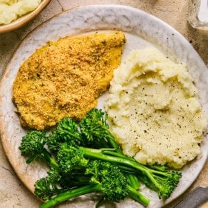 A plate with baked chicken cutlets, a serving of mashed potatoes topped with black pepper, and a side of steamed broccolini. A fork and knife are set beside the plate, while a bowl of creamy mashed potatoes and a salt shaker complete the cozy setup.