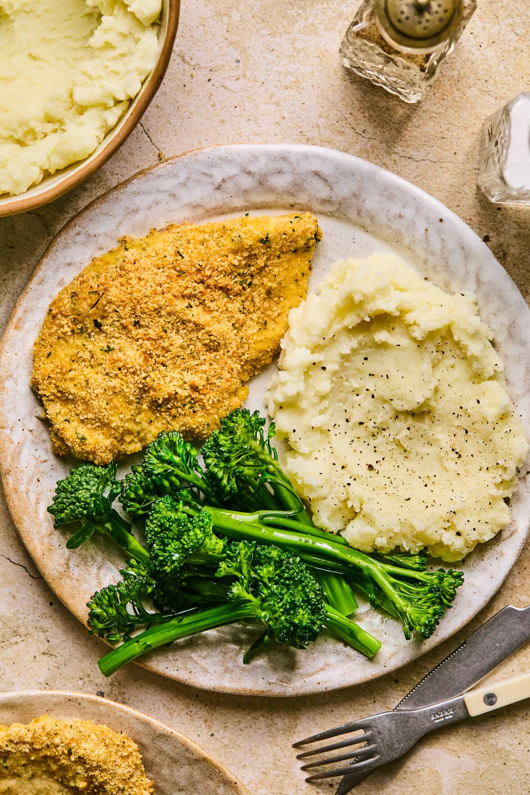 A plate with baked chicken cutlets, a serving of mashed potatoes topped with black pepper, and a side of steamed broccolini. A fork and knife are set beside the plate, while a bowl of creamy mashed potatoes and a salt shaker complete the cozy setup.