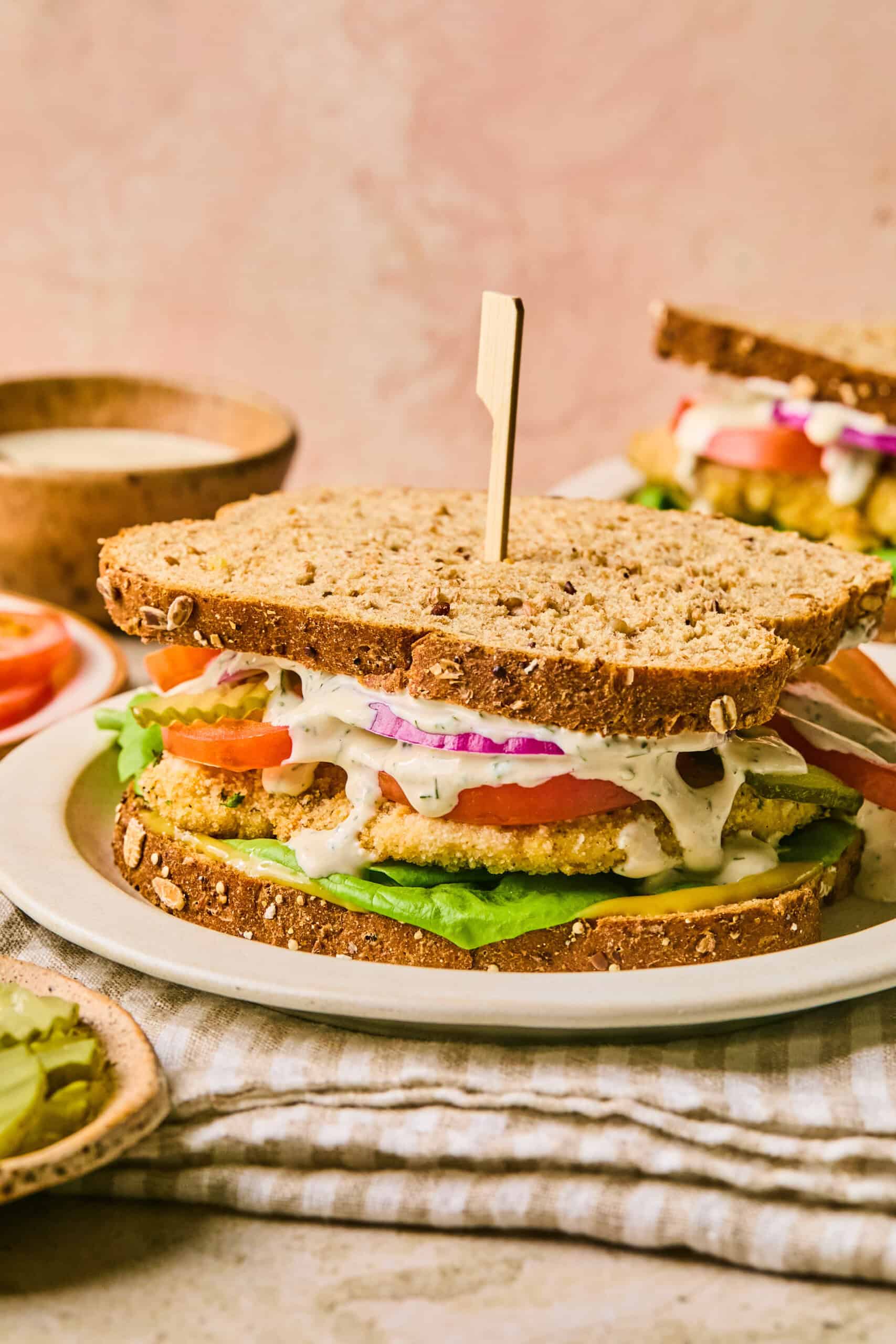 A chicken cutlet sandwich with whole-grain bread, layered with lettuce, tomato slices, red onion, and a crispy fried patty, drizzled with sauce. It is held together by a wooden skewer and placed on a white plate with a striped napkin underneath.