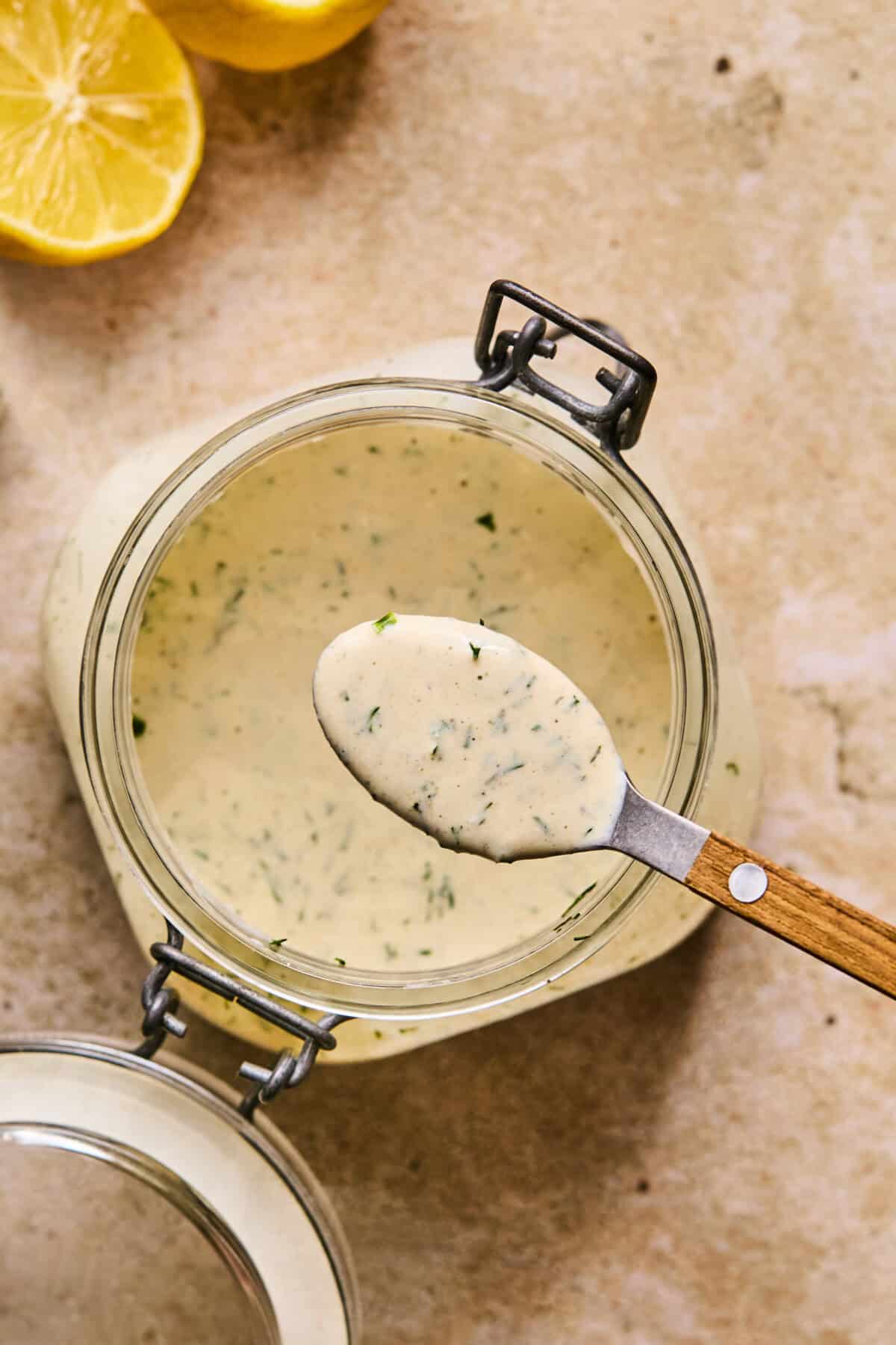 A glass jar filled with creamy, dairy-free ranch dressing sits on a textured surface. A spoon rests on top, revealing the dressings thick consistency and visible herbs. Lemon slices are partially visible in the top left corner, adding a hint of zesty charm.