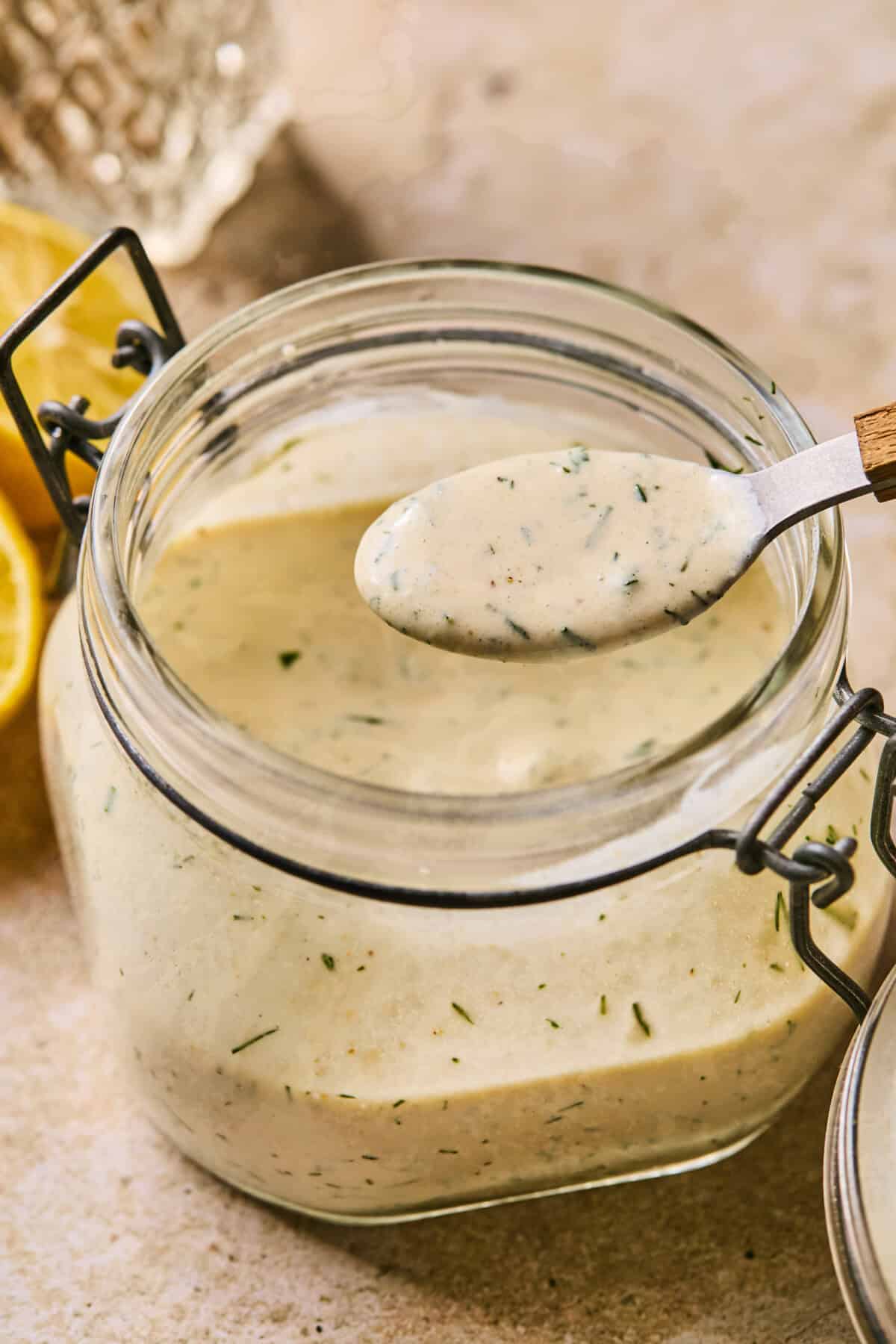 A glass jar filled with creamy, dairy-free ranch dressing, speckled with herbs. A spoon with a dollop of dressing hovers above the open jar. Lemon halves are visible in the background on a textured surface.