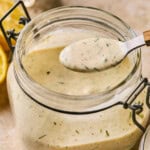 A glass jar filled with creamy, dairy-free ranch dressing, speckled with herbs. A spoon with a dollop of dressing hovers above the open jar. Lemon halves are visible in the background on a textured surface.