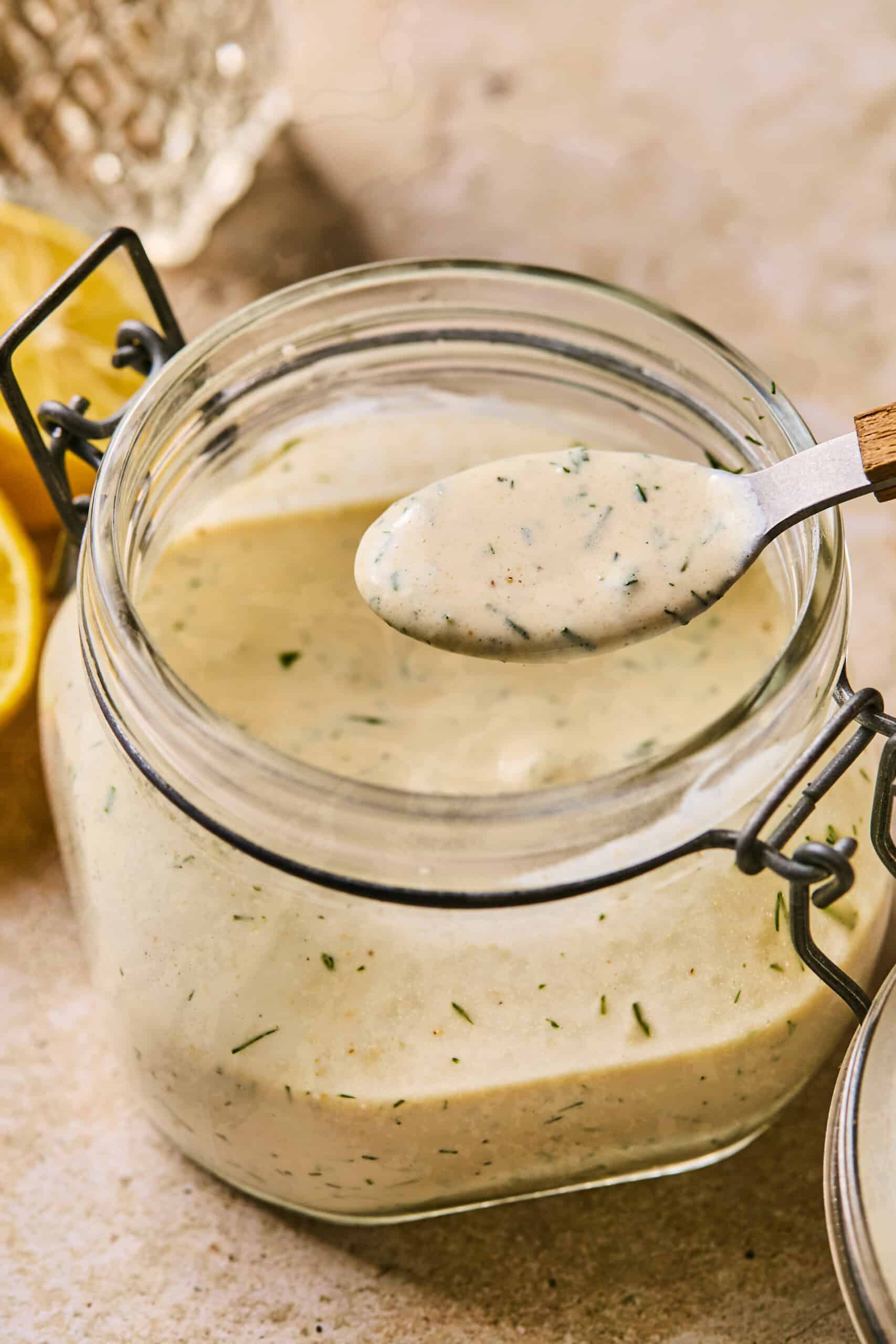 A glass jar filled with creamy, dairy-free ranch dressing, speckled with herbs. A spoon with a dollop of dressing hovers above the open jar. Lemon halves are visible in the background on a textured surface.