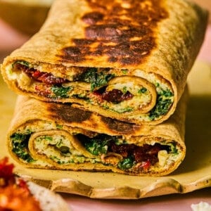 Two grilled spinach and feta wraps from Starbucks are cut in half and stacked. They are filled with greens, sun-dried tomatoes, and cheese, displayed on a wooden board against a pink surface.