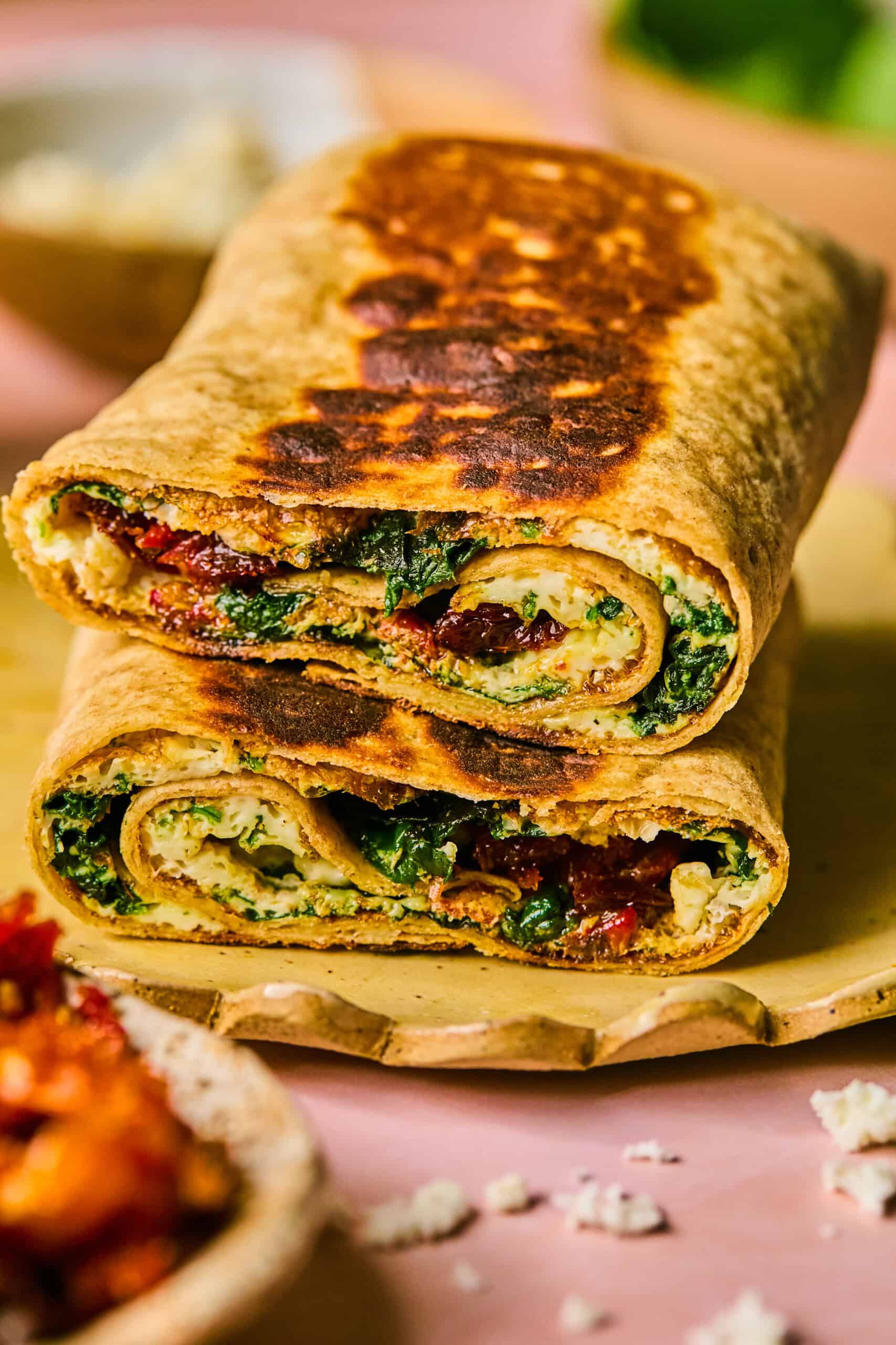 Two grilled spinach and feta wraps from Starbucks are cut in half and stacked. They are filled with greens, sun-dried tomatoes, and cheese, displayed on a wooden board against a pink surface.