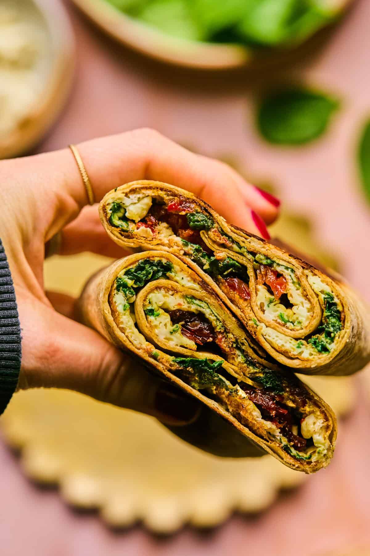 Close-up of a hand holding a sliced spinach and feta wrap filled with greens, tomatoes, and other vegetables. The tightly rolled wrap is displayed against a blurred background reminiscent of the inviting atmosphere at Starbucks, complete with a small plate and scattered greens.