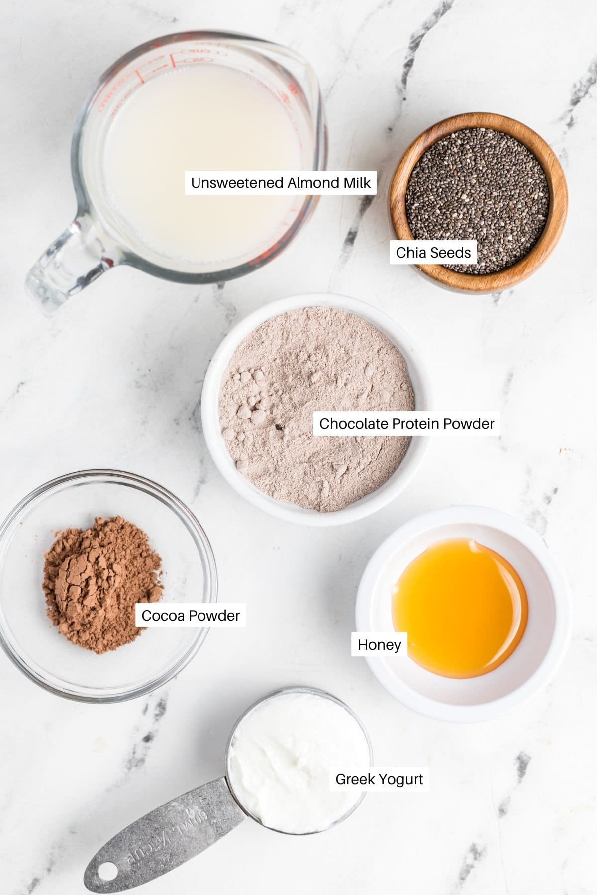 A white surface showcases the makings of a delectable protein chia seed pudding: unsweetened almond milk in a measuring cup, chia seeds in a wooden bowl, chocolate protein powder, cocoa powder, honey, and Greek yogurt all artfully arranged in their own containers.