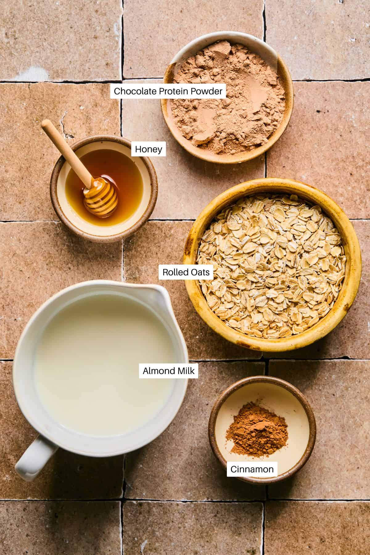 Ingredients arranged on a tiled surface: chocolate protein powder in a bowl, honey in a small bowl with a honey dipper, rolled oats ready for protein oatmeal in a large bowl, a jug of almond milk, and a bowl of cinnamon.