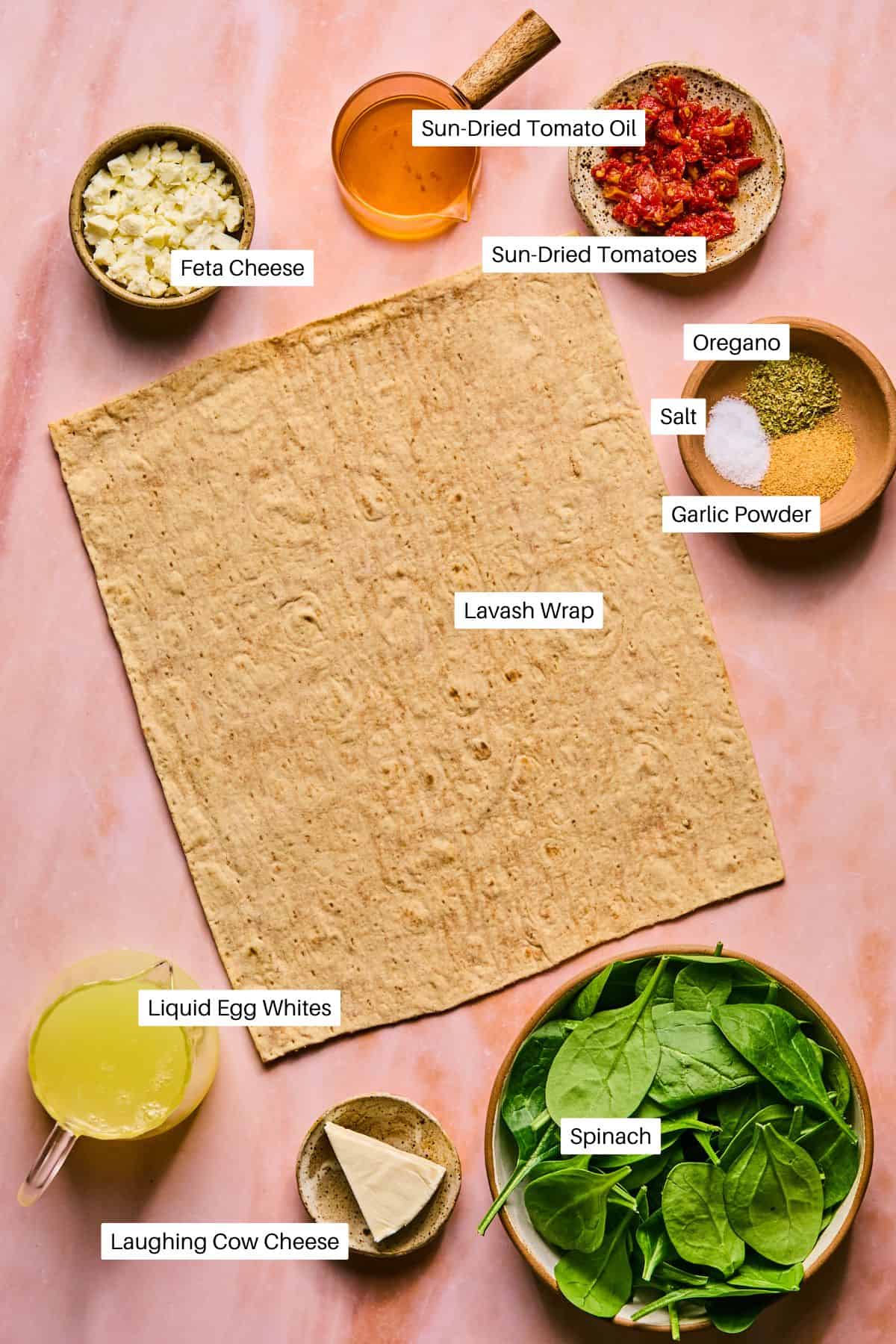 On a pink surface, prepare a delicious spinach and feta wrap inspired by Starbucks. Layer lavash with feta cheese, sun-dried tomatoes in oil, oregano, salt, garlic powder, liquid egg whites, Laughing Cow cheese, and fresh spinach for a delightful meal.