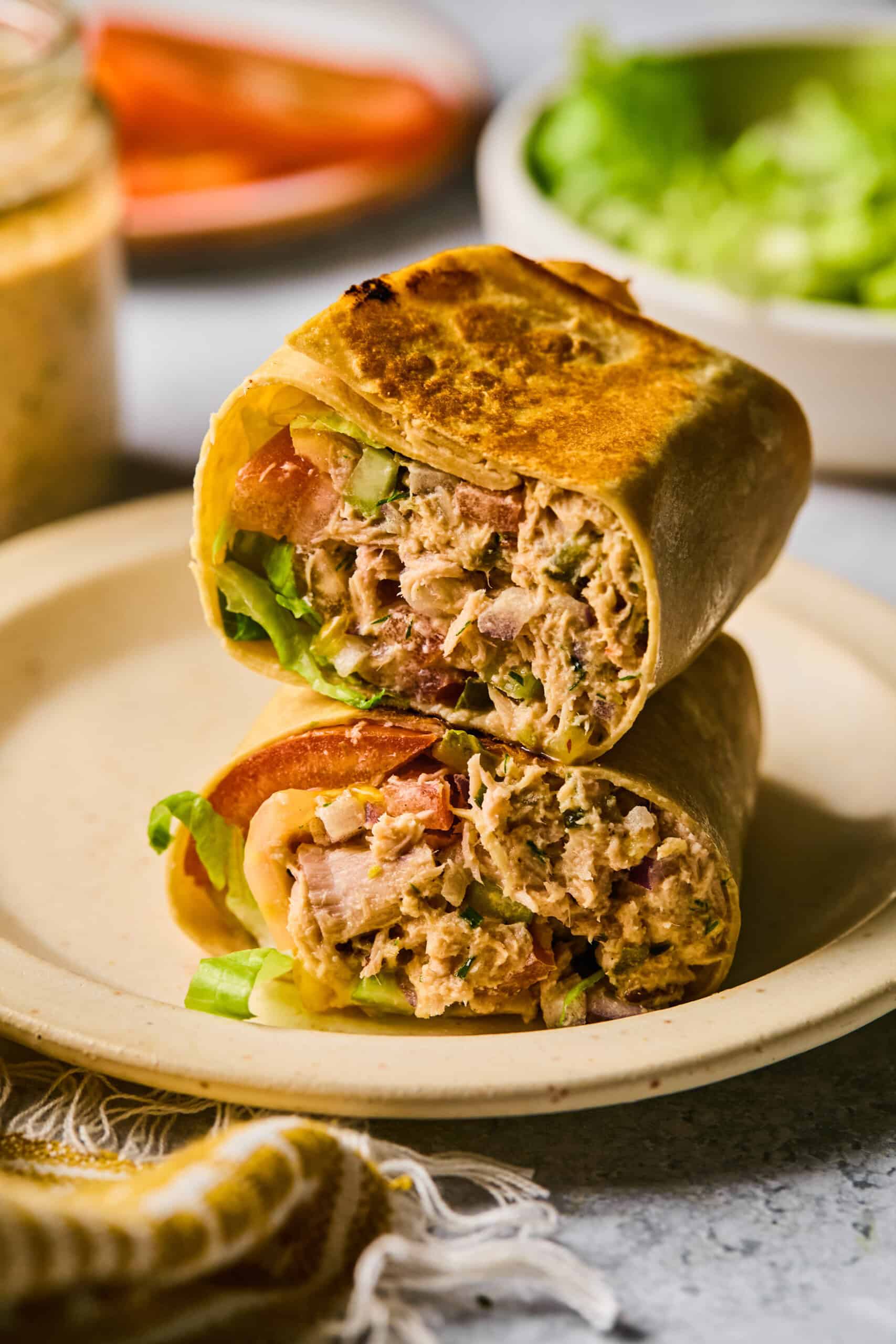 A grilled tuna wrap, cut in half and stacked, is filled with fresh lettuce, juicy tomato, and diced vegetables. It sits invitingly on a beige plate against a blurred background of lettuce, a jar of sauce, and part of a pepper. A cloth napkin is partially visible.