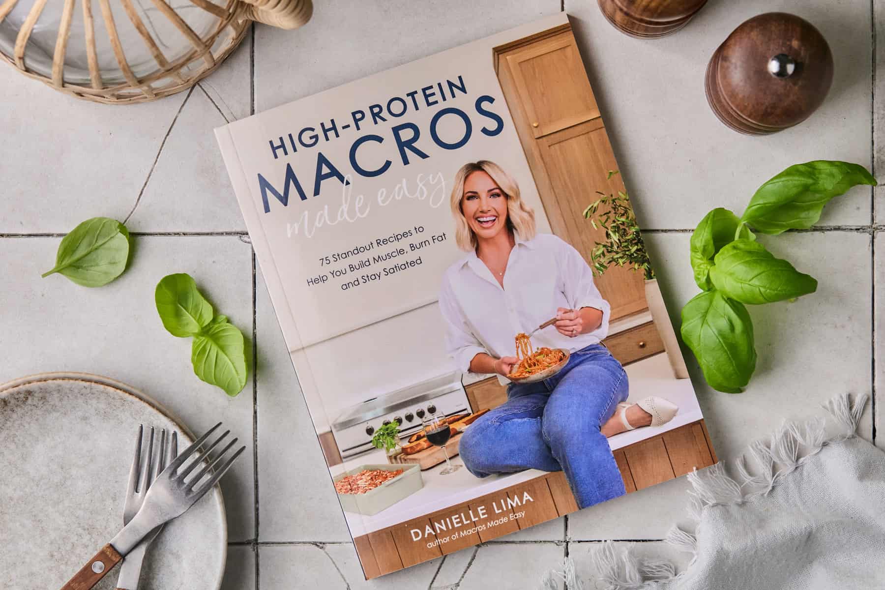 A cookbook titled High-Protein Macros Made Easy by Danielle Lima is on a tiled surface. The cover features a smiling woman sitting in a kitchen, eating spaghetti. Surrounding items include greens, a fork, knife, and wooden pepper mills.