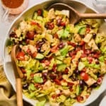 A large bowl of Mediterranean chopped salad brimming with lettuce, cherry tomatoes, cucumber, olives, feta cheese, red beans, and grilled chicken pieces. Wooden serving spoons rest inside the bowl, with dressing and salt and pepper shakers nearby.