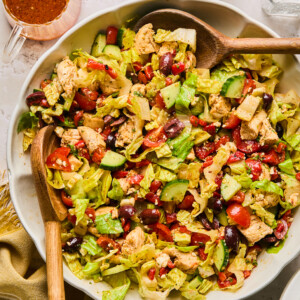 A large bowl of Mediterranean chopped salad brimming with lettuce, cherry tomatoes, cucumber, olives, feta cheese, red beans, and grilled chicken pieces. Wooden serving spoons rest inside the bowl, with dressing and salt and pepper shakers nearby.