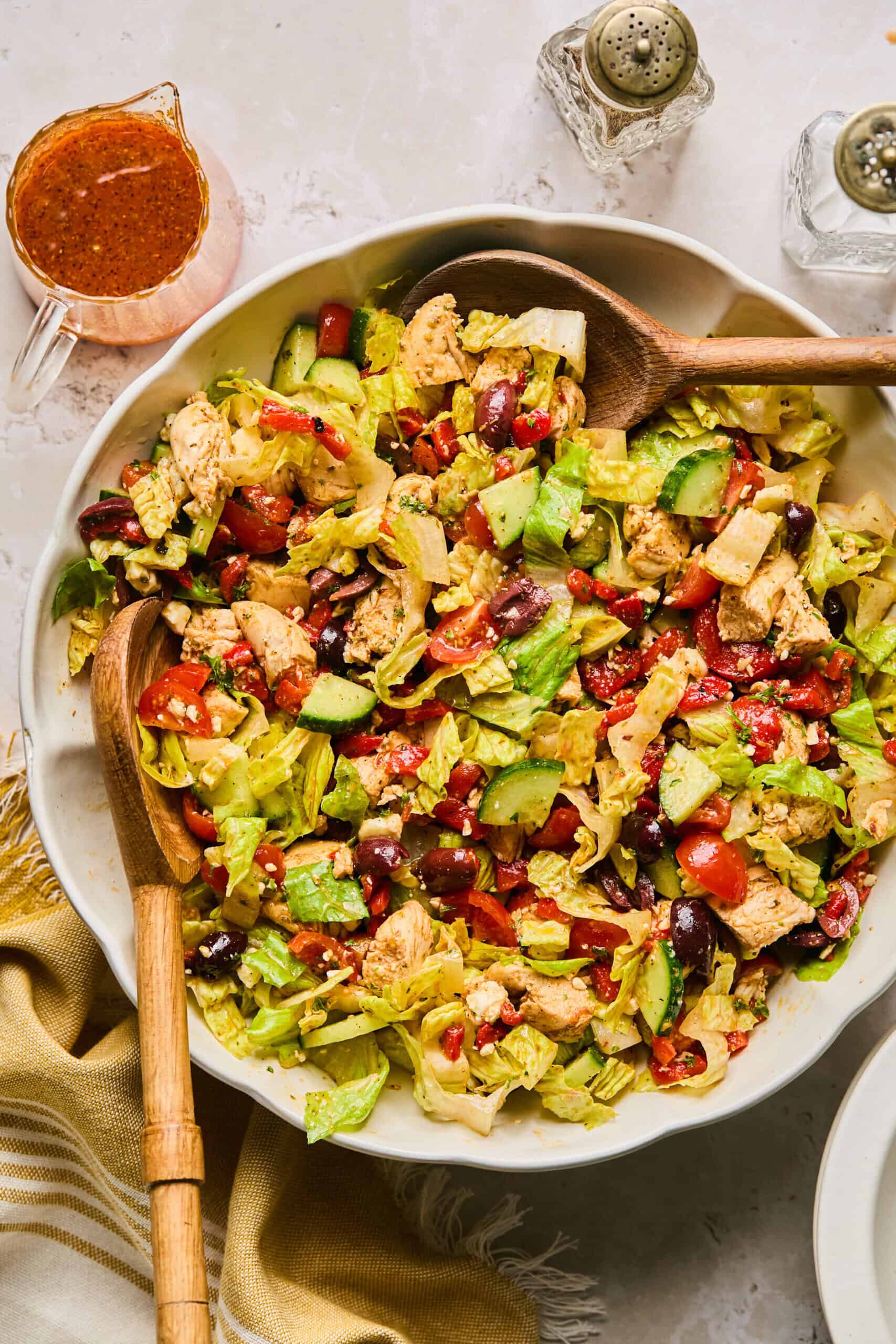 A large bowl of Mediterranean chopped salad brimming with lettuce, cherry tomatoes, cucumber, olives, feta cheese, red beans, and grilled chicken pieces. Wooden serving spoons rest inside the bowl, with dressing and salt and pepper shakers nearby.