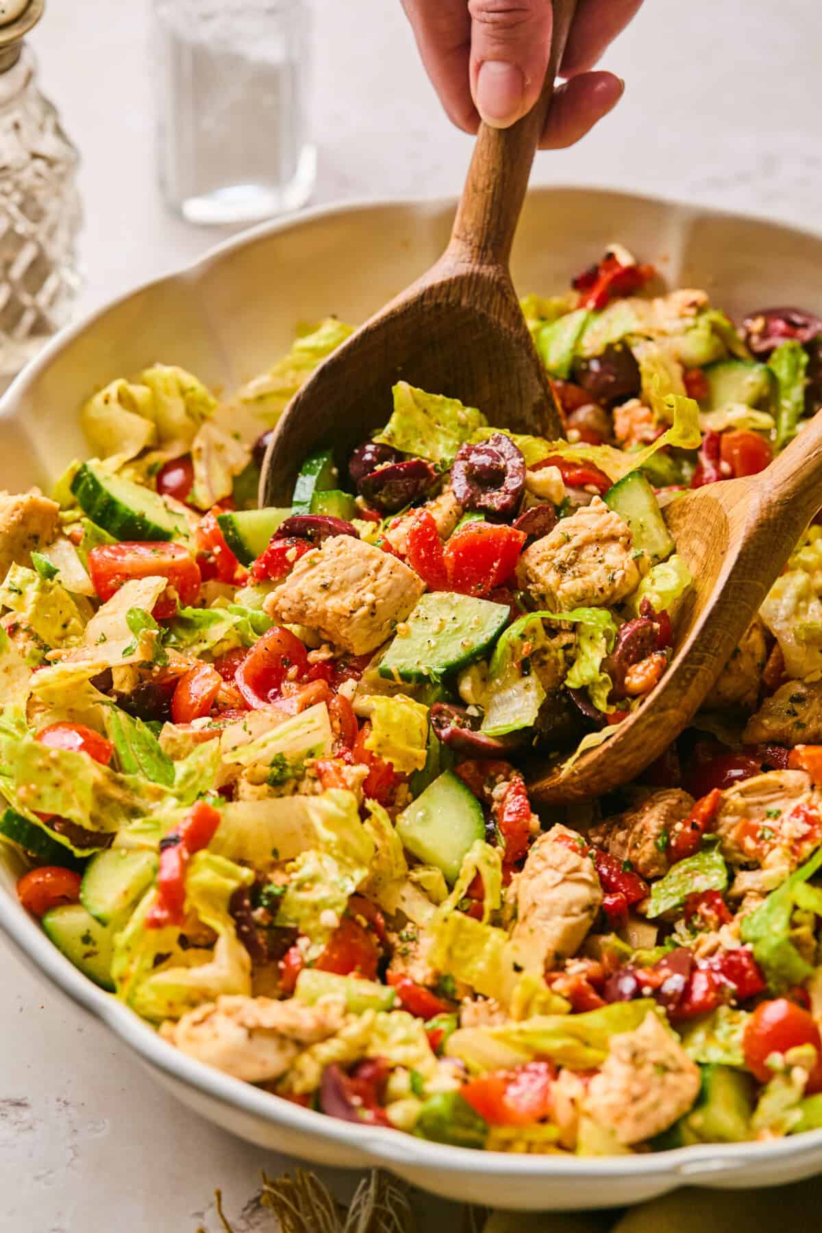 A vibrant Mediterranean chopped salad with lettuce, cucumbers, tomatoes, olives, and chunks of grilled chicken is tossed with wooden salad servers. A hand is visible holding one of the servers.