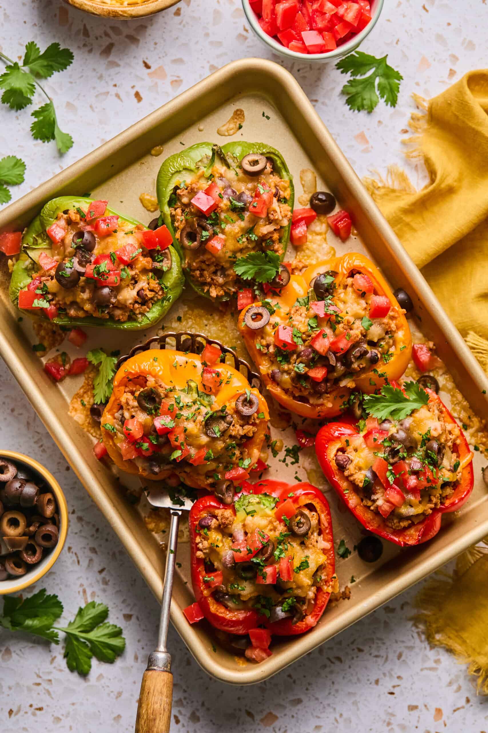 A baking tray brimming with colorful taco stuffed peppers, crowned with melted cheese, tomatoes, and olives. The peppers are garnished with parsley. Nearby, small bowls of chopped tomatoes and olives accompany a cheerful yellow napkin.