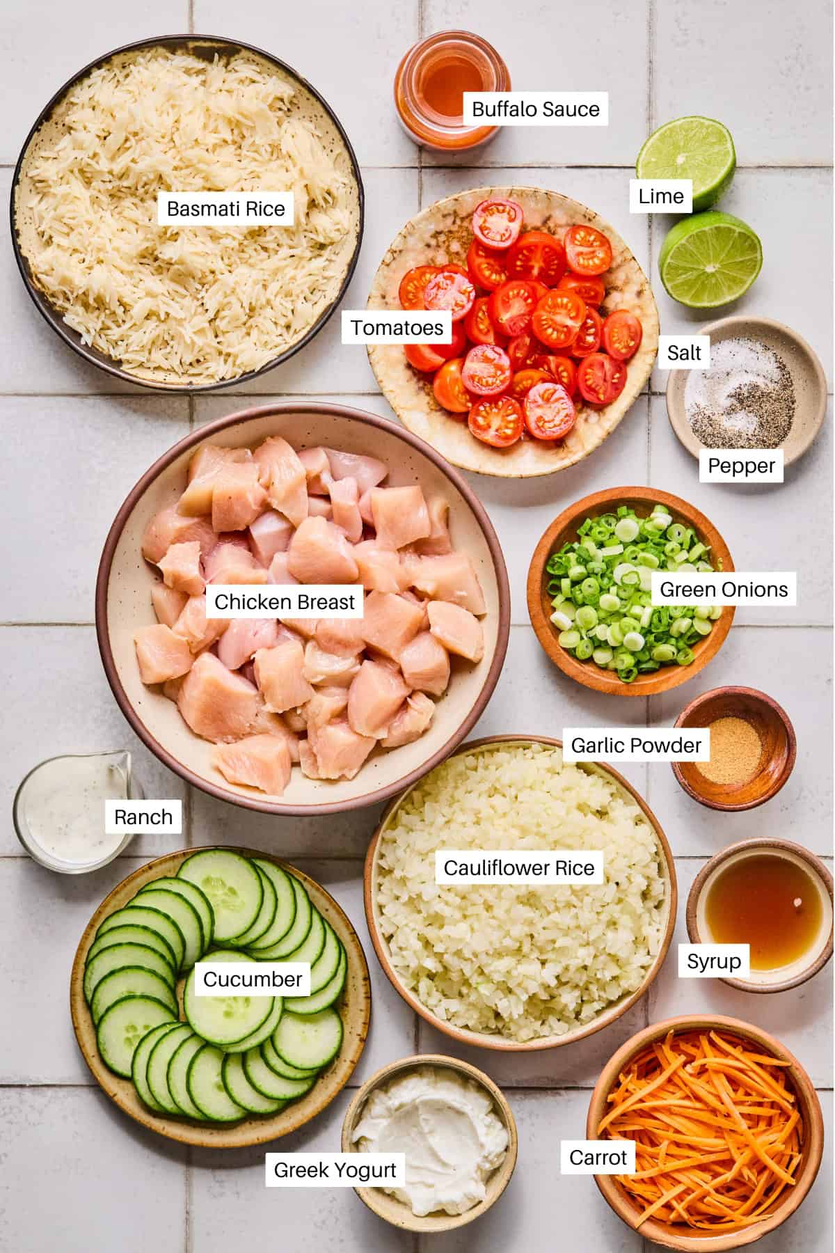 A top-down view of a Buffalo Chicken Rice Bowl featuring basmati rice, chicken breast pieces tossed in buffalo sauce, lime, salt, pepper, tomatoes, green onions, garlic powder, ranch, cucumber, cauliflower rice, syrup, Greek yogurt, and carrot.