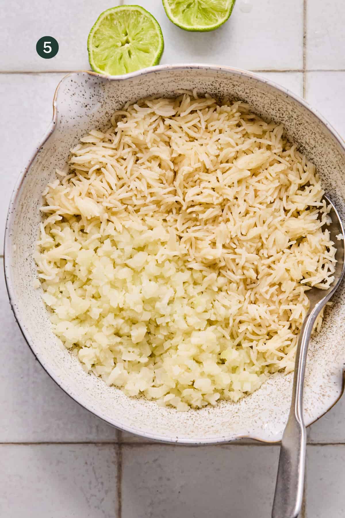 A bowl containing cooked white rice and cauliflower rice, side by side, with a spoon resting inside—perfect for a Buffalo Chicken Rice Bowl. Two lime halves are visible at the top of the image on a white tiled surface.