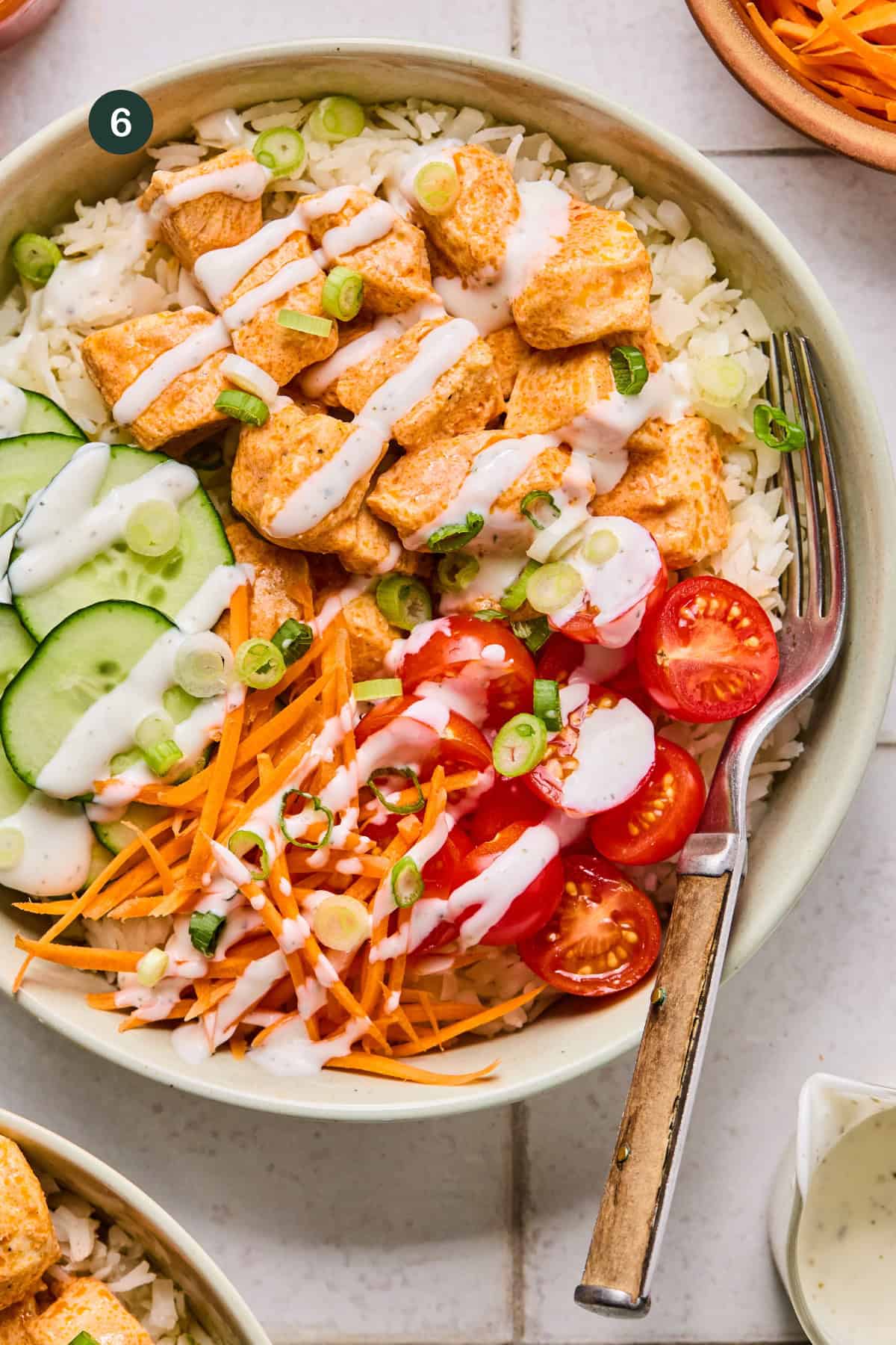 A Buffalo Chicken Rice Bowl filled with rice, seasoned chicken pieces, sliced cucumbers, shredded carrots, cherry tomato halves, and chopped green onions is drizzled with creamy white dressing. A fork rests inside the bowl.
