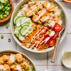 A Buffalo Chicken Rice Bowl filled with rice, sliced cucumber, shredded carrots, cherry tomatoes, seasoned chicken pieces, and green onions is drizzled with creamy dressing, surrounded by similar bowls and toppings on a white tiled surface.