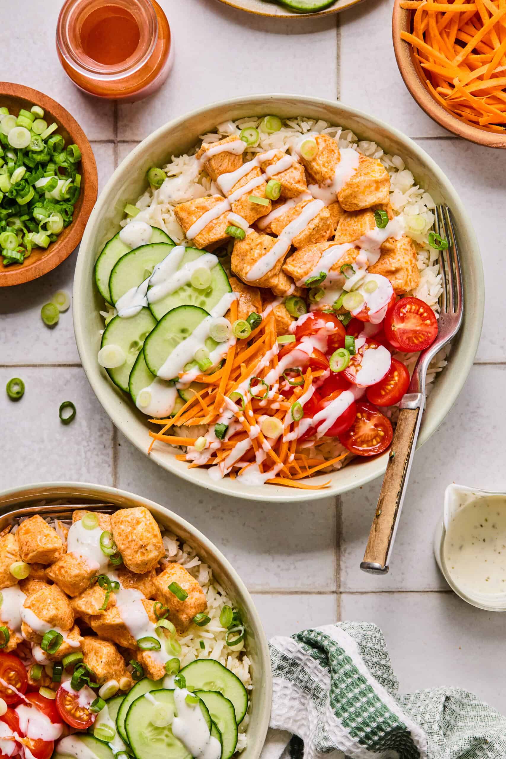 A Buffalo Chicken Rice Bowl filled with rice, sliced cucumber, shredded carrots, cherry tomatoes, seasoned chicken pieces, and green onions is drizzled with creamy dressing, surrounded by similar bowls and toppings on a white tiled surface.