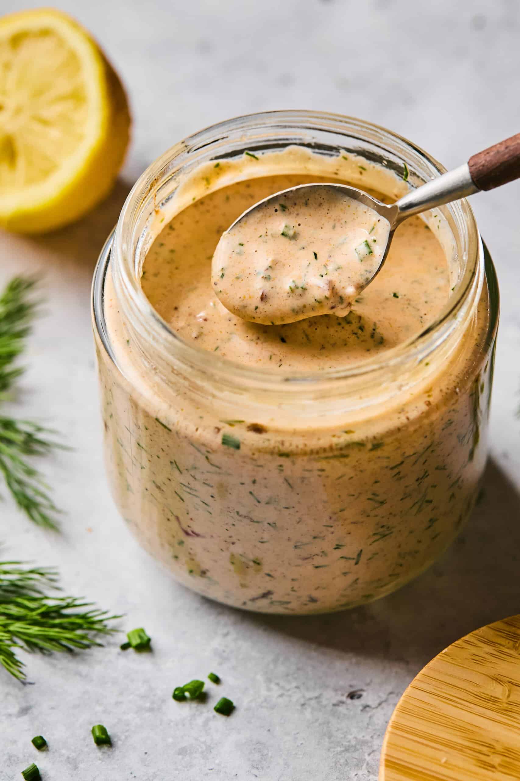 A glass jar filled with creamy chipotle ranch dressing, speckled with herbs and spices, sits on a light surface. A spoon rests above the jar, dipping into the sauce. Fresh dill, chopped chives, and a halved lemon are placed nearby.