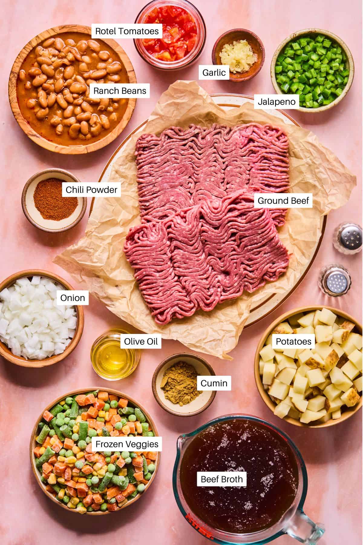 Flat lay of cowboy soup ingredients on a pink surface: ground beef, diced potatoes, frozen mixed veggies, chopped onion, ranch beans, Rotel tomatoes, garlic, jalapeño, chili powder, cumin, olive oil, and beef broth.