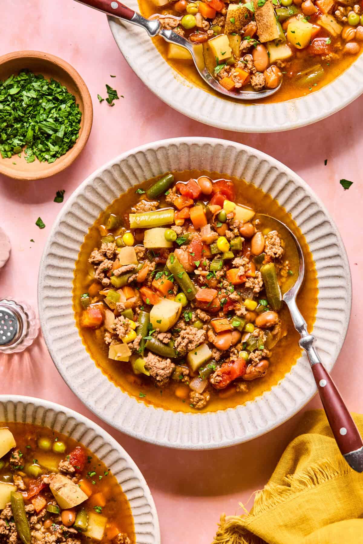 A bowl of hearty cowboy soup with ground beef, potatoes, green beans, corn, peas, carrots, pinto beans, and tomatoes, garnished with parsley. The bowl sits on a pink surface beside other bowls and a small dish of chopped herbs.