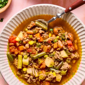 A bowl of hearty cowboy soup with ground meat, green beans, carrots, tomatoes, potatoes, peas, and beans. A spoon rests inside the bowl, and chopped parsley is visible in a small dish nearby.