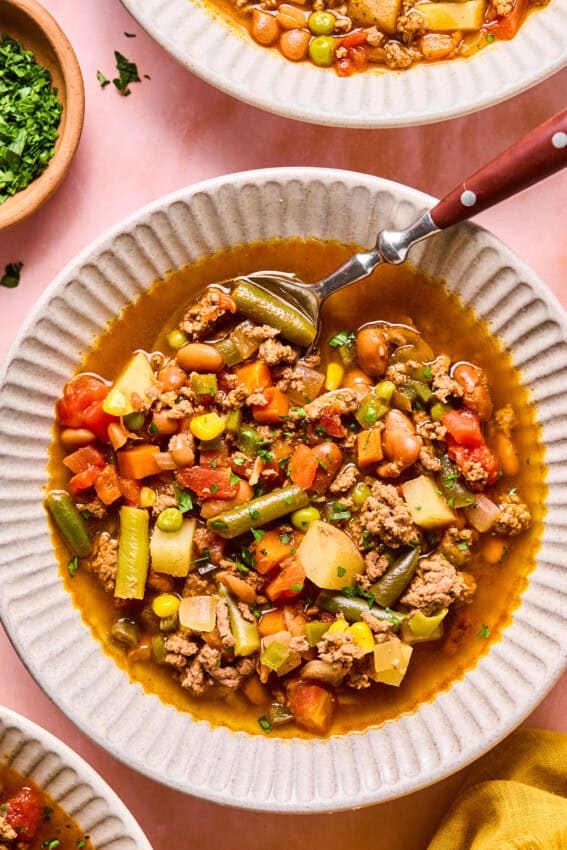 A bowl of hearty cowboy soup with ground meat, green beans, carrots, tomatoes, potatoes, peas, and beans. A spoon rests inside the bowl, and chopped parsley is visible in a small dish nearby.
