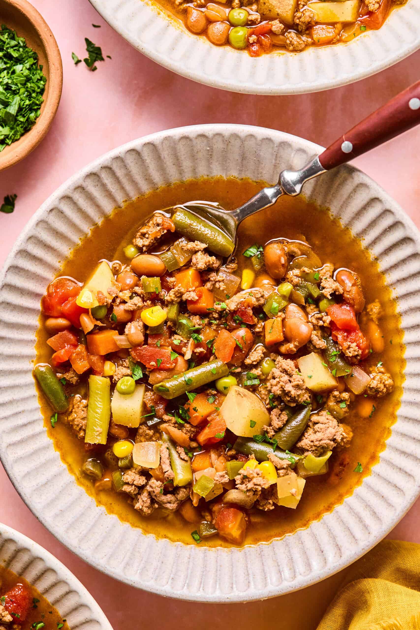 A bowl of hearty cowboy soup with ground meat, green beans, carrots, tomatoes, potatoes, peas, and beans. A spoon rests inside the bowl, and chopped parsley is visible in a small dish nearby.