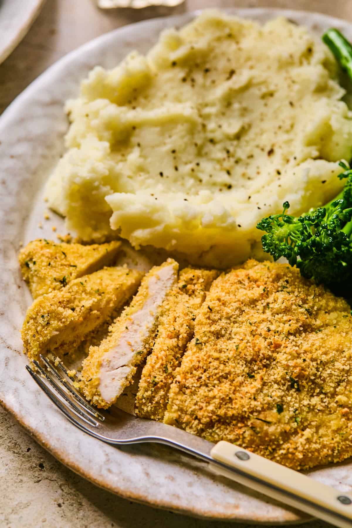 A plate featuring golden baked chicken cutlets next to creamy mashed potatoes and a serving of steamed broccoli, with a fork placed on the side.