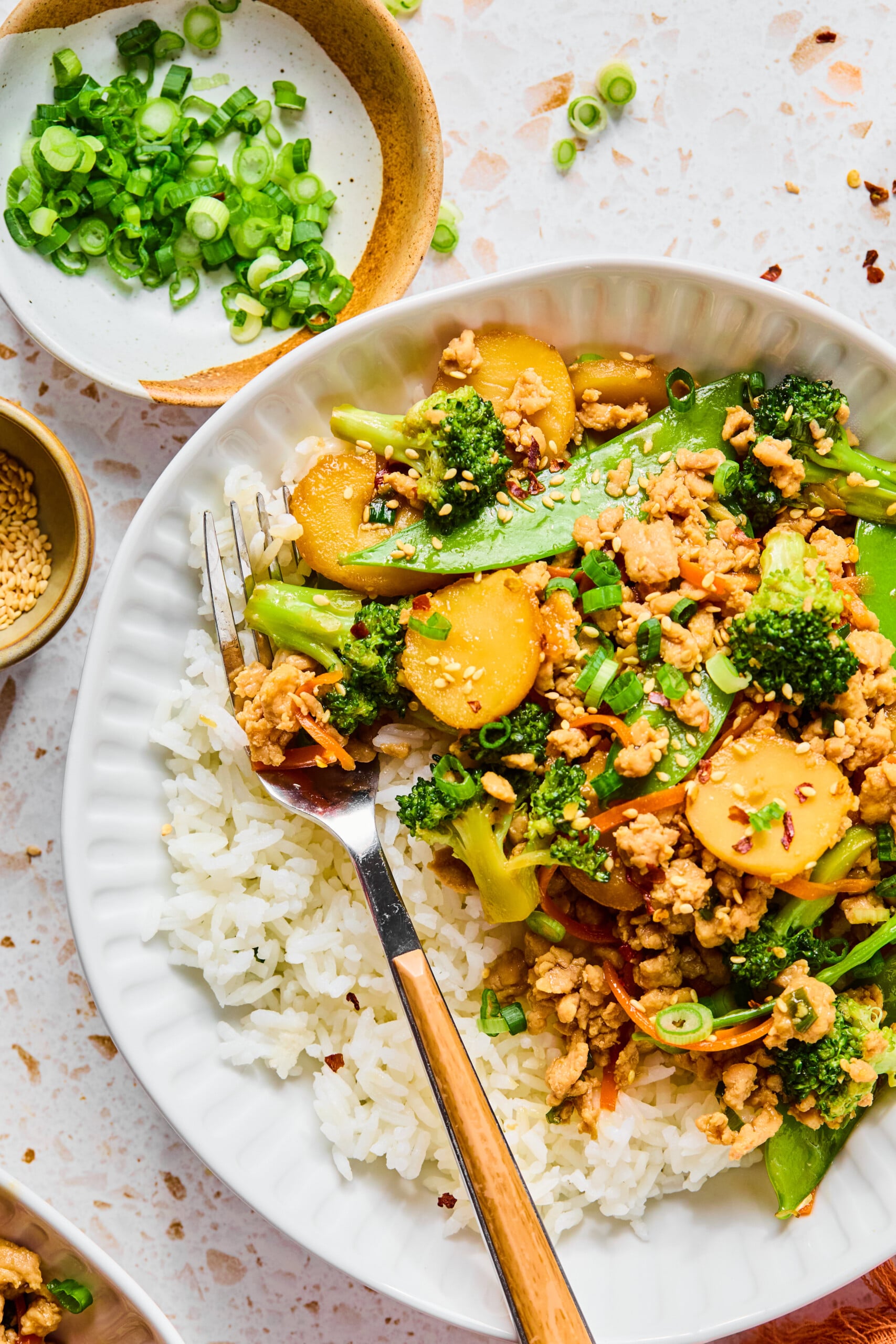 A white plate with white rice and Ground Chicken Stir Fry, featuring broccoli, water chestnuts, carrots, and snap peas. A fork rests on the plate. Diced green onions and sesame seeds are in small bowls nearby.