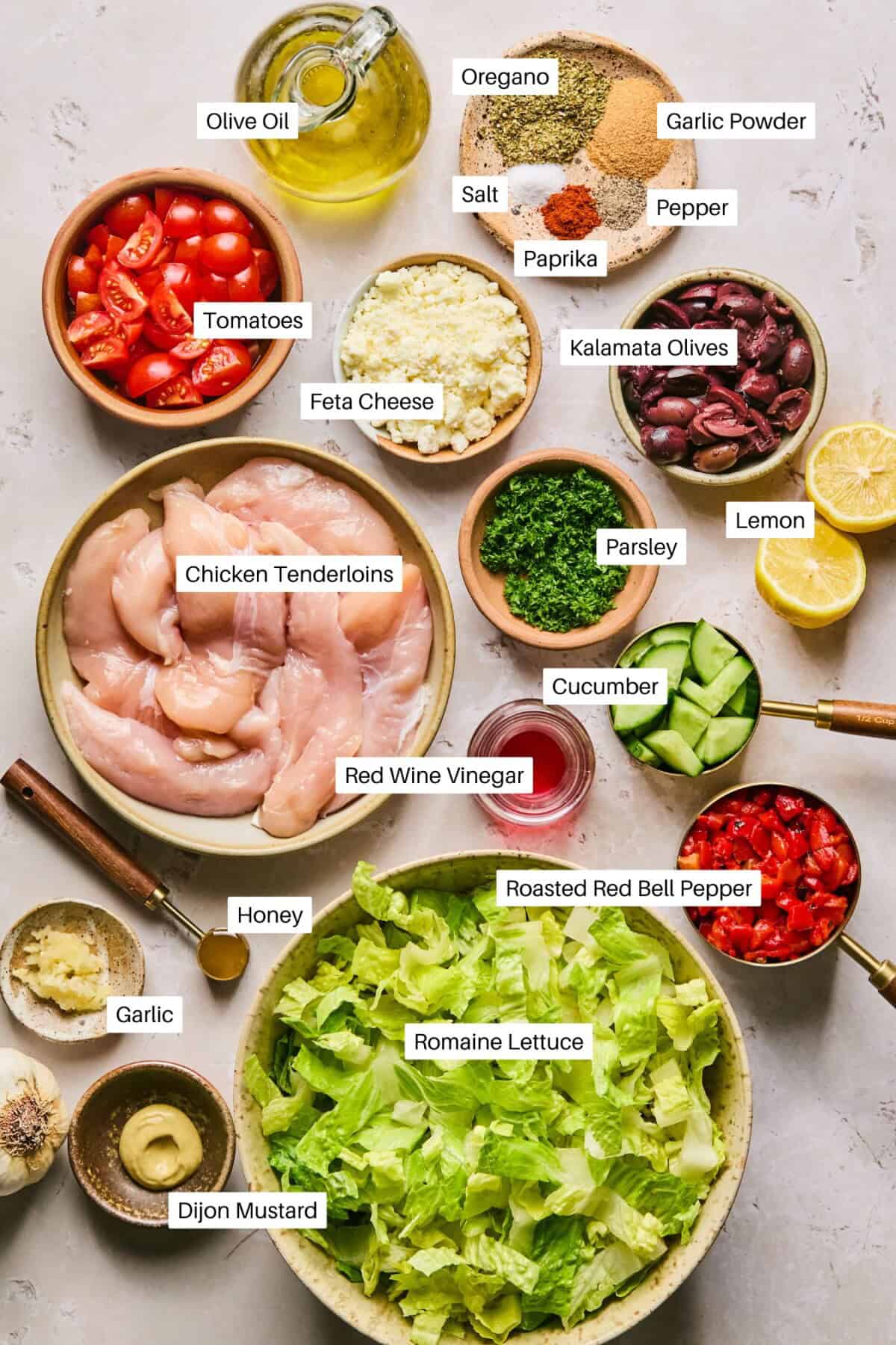 A colorful assortment of ingredients for a Mediterranean chopped salad is labeled and displayed: chicken tenderloins, Romaine lettuce, tomatoes, cucumbers, feta cheese, Kalamata olives, roasted red bell pepper, and various spices and dressings.