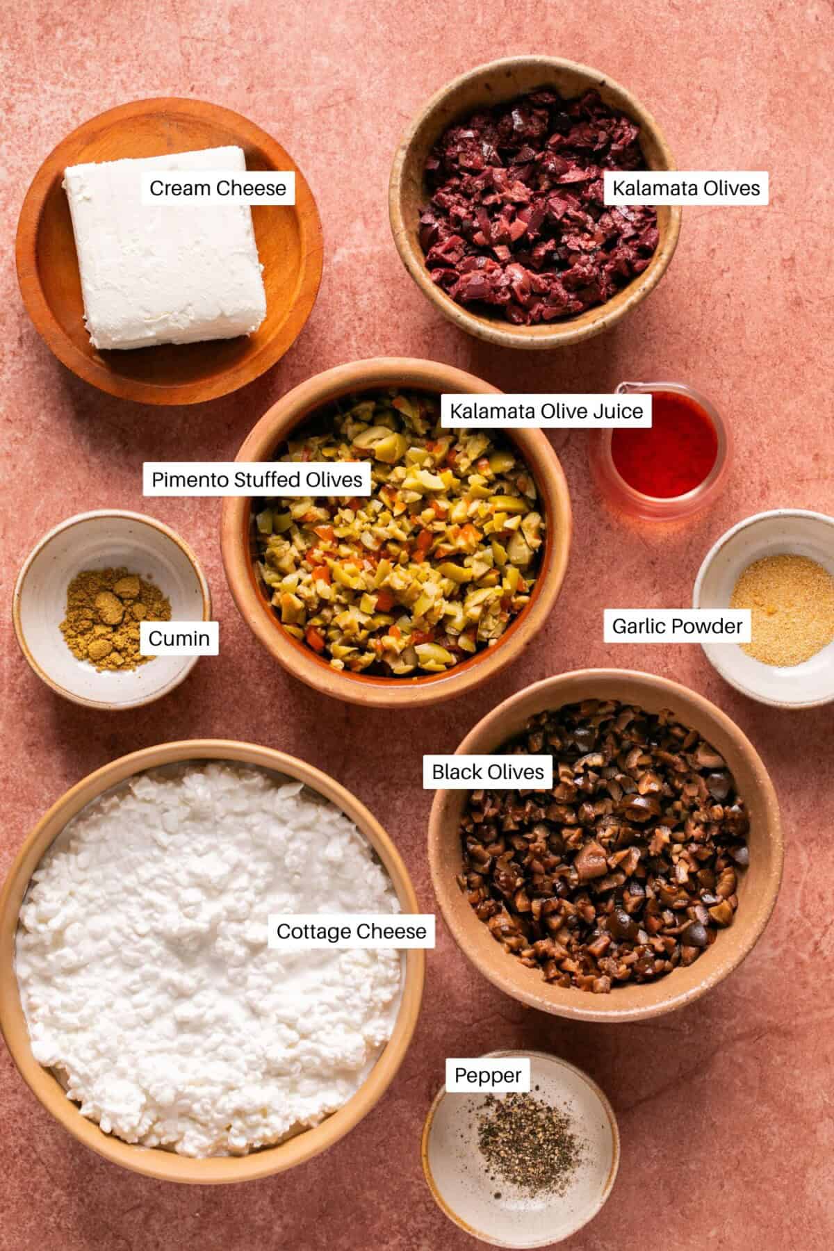 A collection of ingredients for a delicious olive dip is spread across a pink surface, featuring cream cheese, kalamata olives and juice, pimento-stuffed olives, garlic powder, cumin, black olives, cottage cheese, and pepper.
