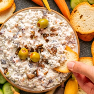 A hand dips a toasted slice of baguette into a bowl of luscious olive dip, rich with creamy texture and topped with olives and chopped veggies. The bowl is surrounded by vibrant carrot sticks, cucumber slices, and extra toasted baguette slices on a sleek gray surface.