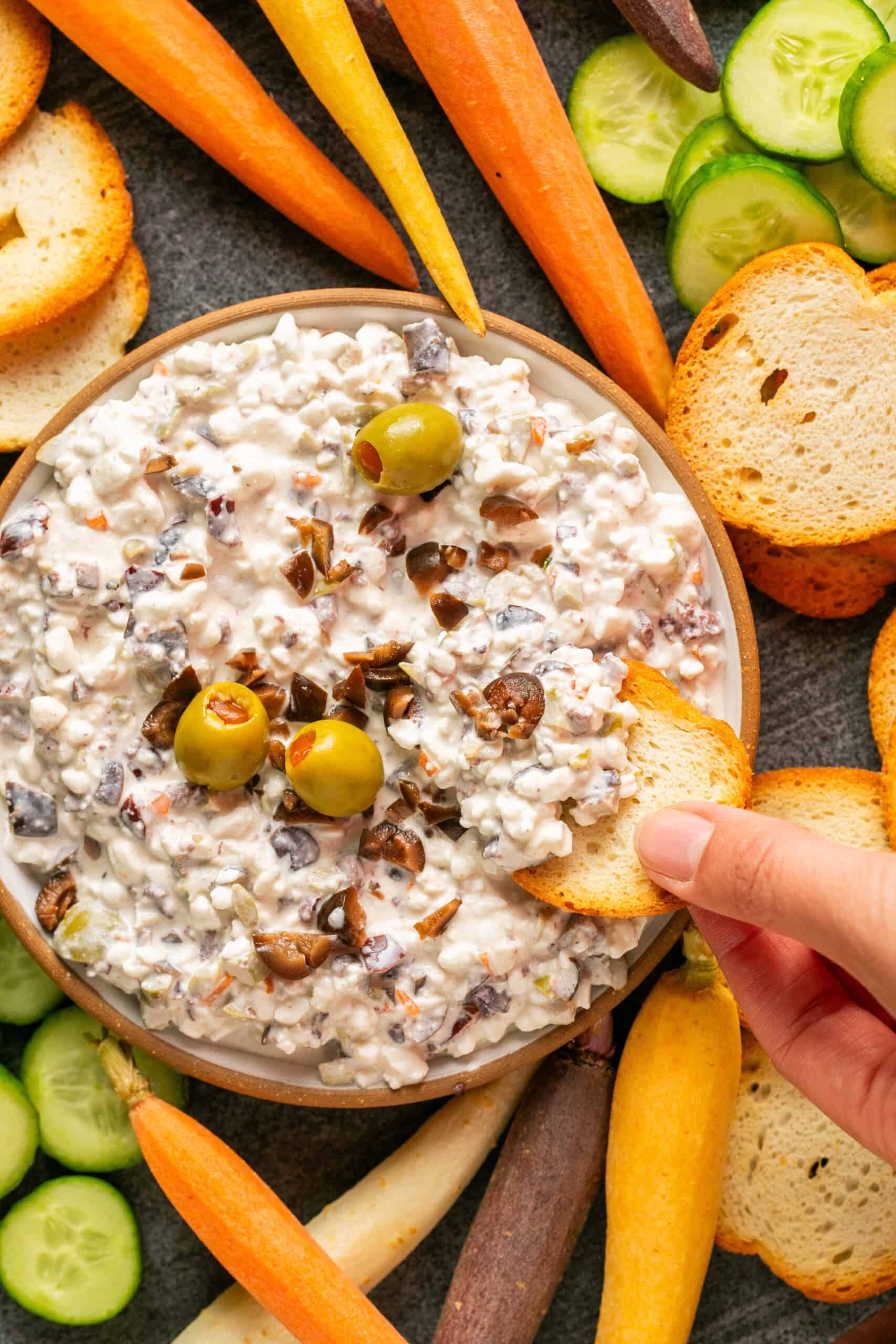 A hand dips a toasted slice of baguette into a bowl of luscious olive dip, rich with creamy texture and topped with olives and chopped veggies. The bowl is surrounded by vibrant carrot sticks, cucumber slices, and extra toasted baguette slices on a sleek gray surface.
