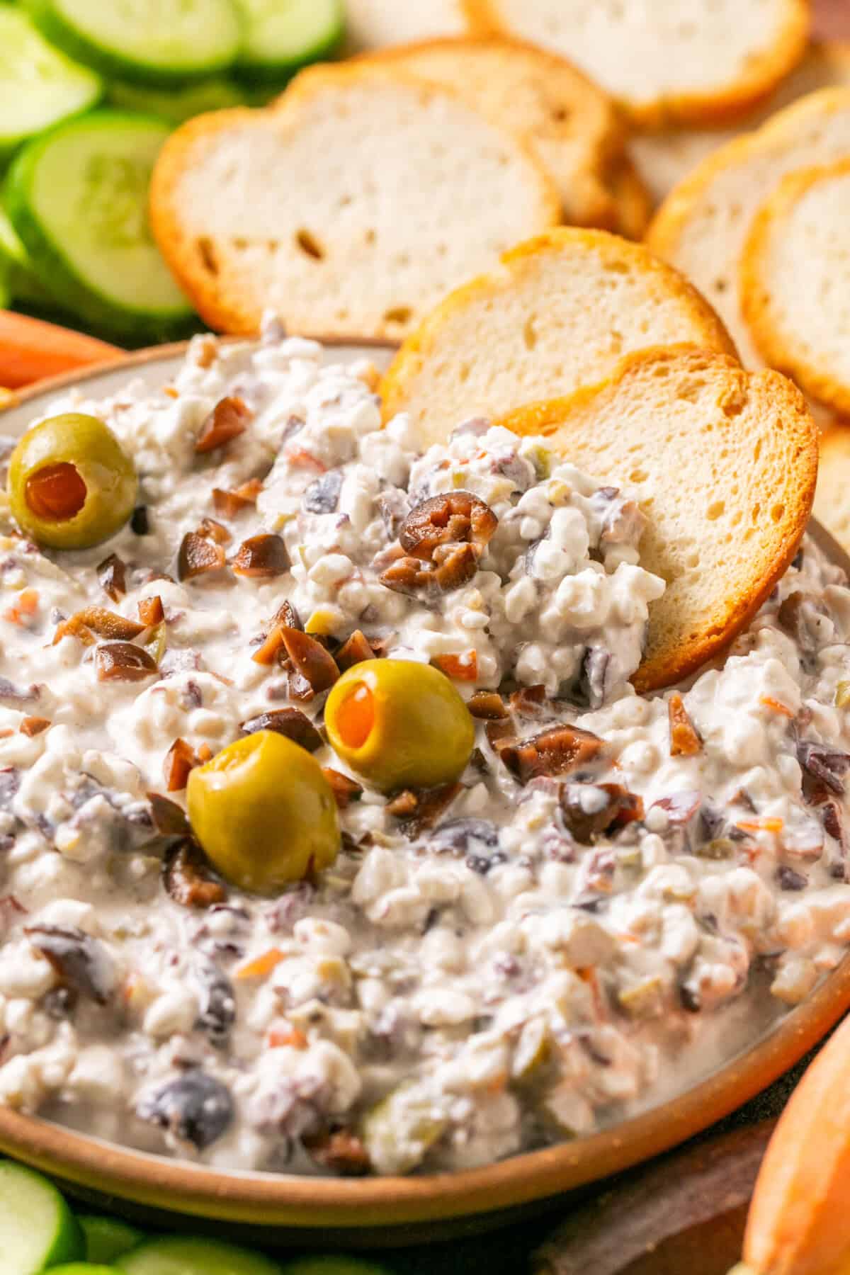 A bowl of creamy olive dip with chopped olives and whole green olives on top is served alongside slices of toasted bread. Cucumber slices can be seen in the background.