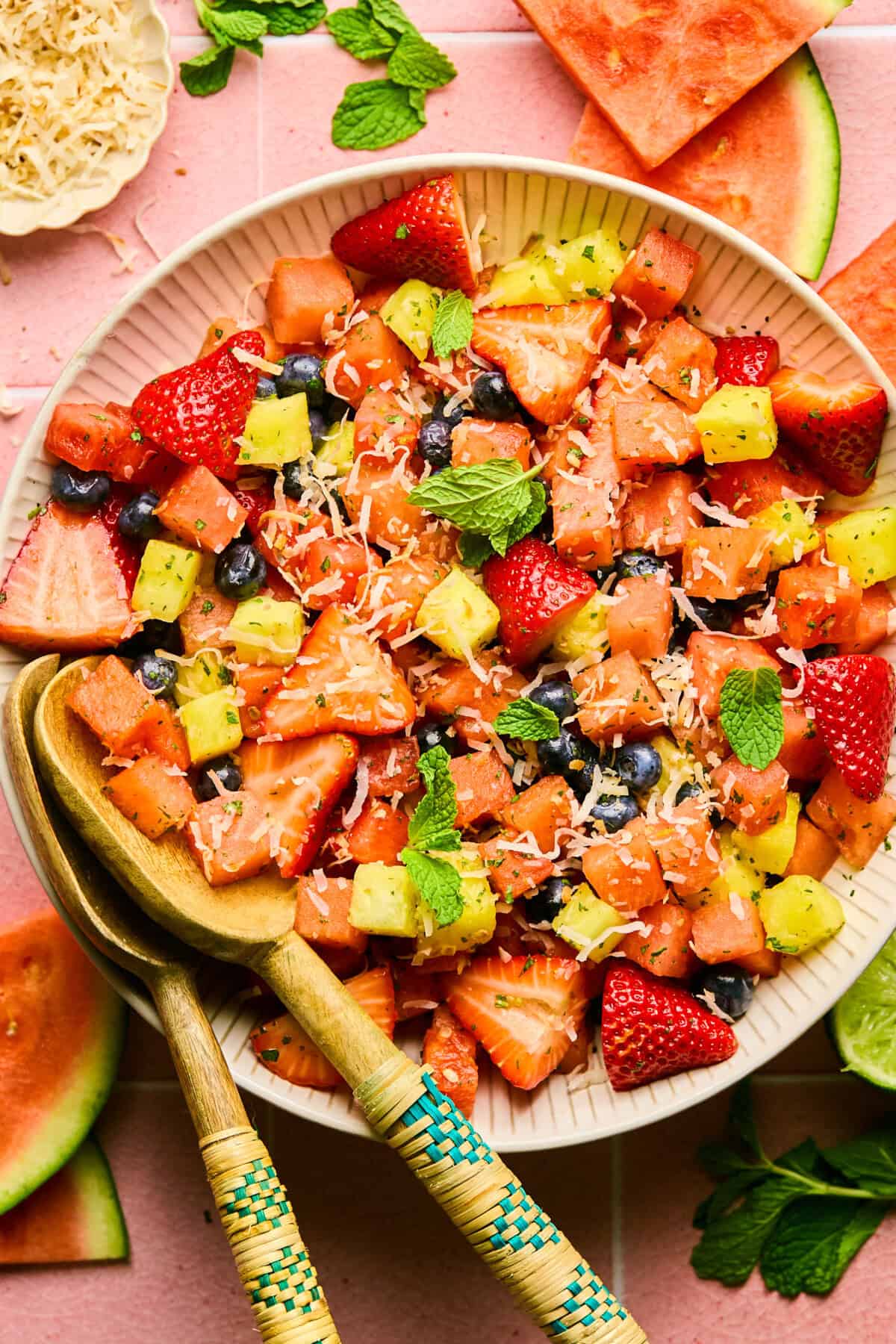 A bowl of vibrant Watermelon Fruit Salad with strawberries, pineapple, blueberries, shredded coconut, and mint leaves. Wooden serving utensils rest in the bowl, and fruit slices decorate the pink tiled surface underneath.